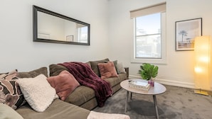 Un salon comprenant un canapé, une table basse, un grand miroir et une fenêtre donnant sur des immeubles.