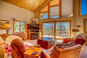 A spacious living room with a fireplace, wooden furniture, and large windows offering a view of the outdoors.