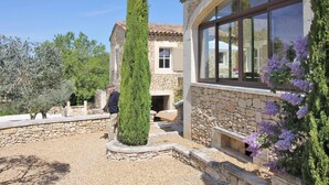 A stone building with a large arched window, a well-maintained garden with gravel paths, and a tall coniferous tree. - Rural bastide and outhouse with private pool (Apt)