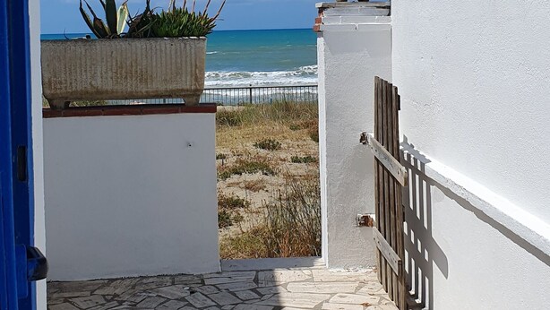 Un sendero pavimentado que lleva a una playa, con una pared blanca y una puerta azul a la izquierda.