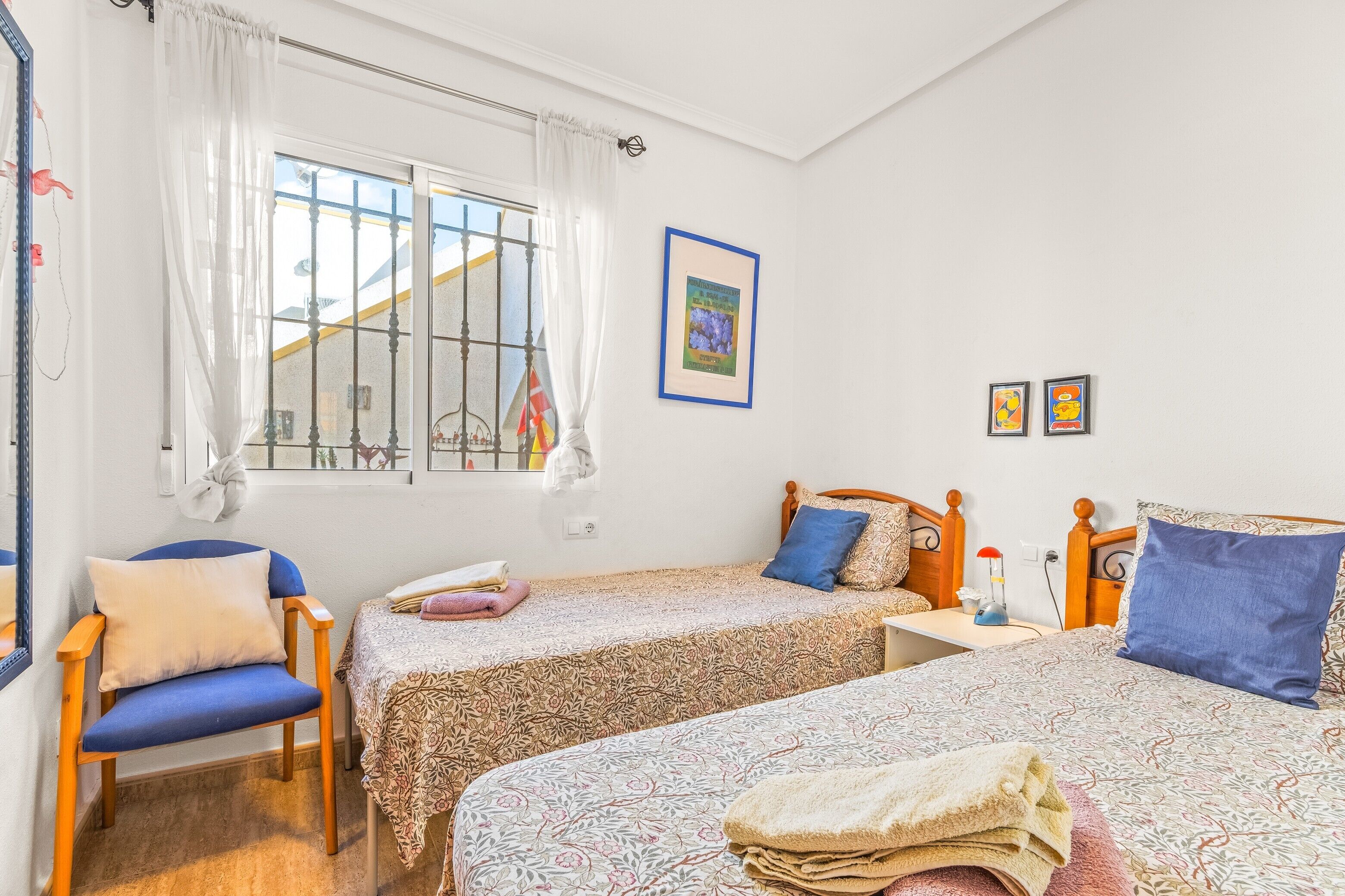 Ein Schlafzimmer mit zwei Betten, einem Sessel, einem Fenster mit Vorhängen und einem gerahmten Bild an der Wand.