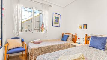 Ein Schlafzimmer mit zwei Betten, einem Sessel, einem Fenster mit Vorhängen und einem gerahmten Bild an der Wand.