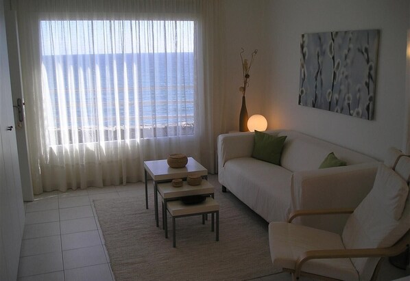 A living room with a sofa, a chair, a table, a lamp, and a view of the sea through sheer curtains.