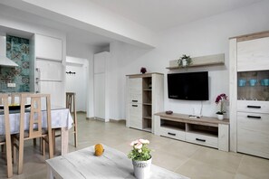 Une cuisine moderne avec des meubles blancs, un coin repas avec des chaises et une table en bois, et un téléviseur fixé au mur.