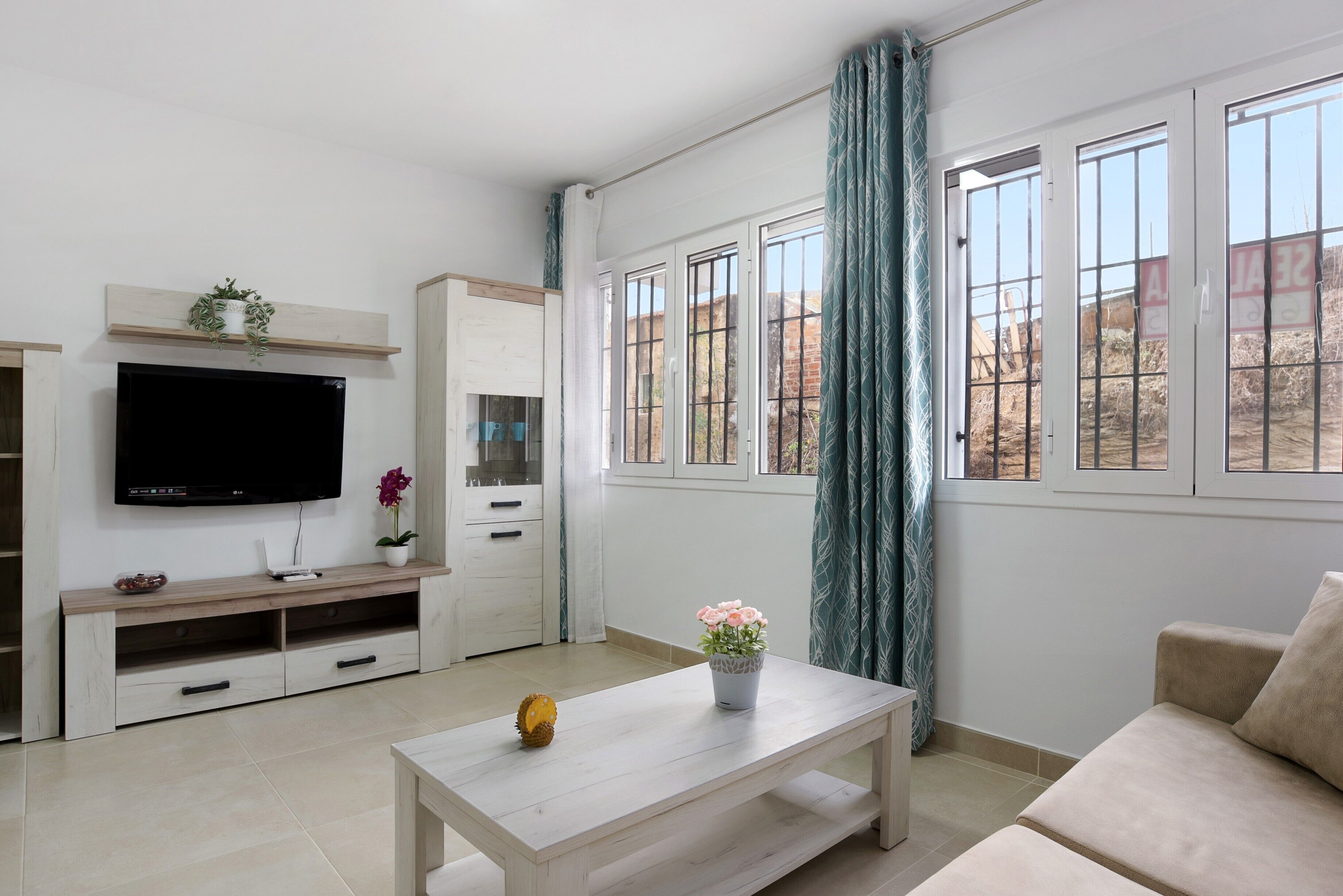 A living room with a TV, a coffee table, a sofa, a window with curtains, and a view of the outside.