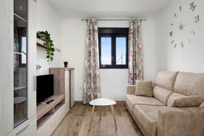 Een moderne woonkamer met een flatscreen televisie, een ronde witte tafel, een beige bank en een raam met bloemen gordijnen.