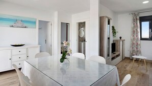 A modern dining area with a glass table, white chairs, a white cabinet with drawers, a painting of a pier, and a television on a wooden stand. - Chalet "Casa Manoli" with Pool, Terrace, Air Conditioning and Wi-Fi (Barrio Nuevo)