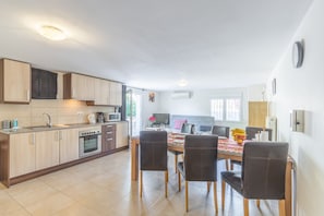 A modern kitchen with wooden cabinets, a dining table with chairs, and a TV in the background.