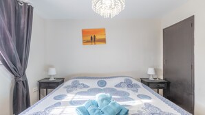 Un dormitorio con una cama grande, dos mesitas de noche con lámparas, una araña, un cuadro de un atardecer y una puerta.
