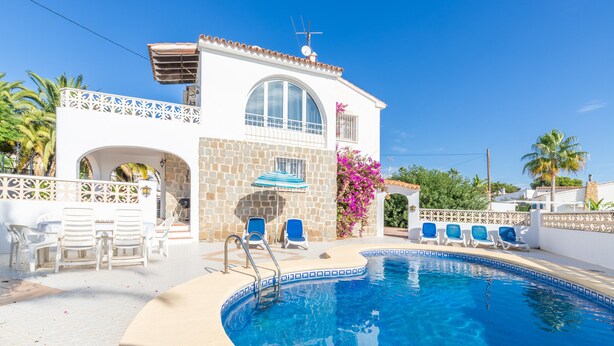 Un área de piscina con un edificio blanco, una zona de estar cubierta y un cielo azul despejado.