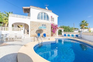 A pool area with a white building, a covered seating area, and a clear blue sky.