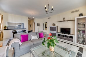 A modern living room with a grey sofa, a glass coffee table, a flat-screen TV, and a kitchen in the background.