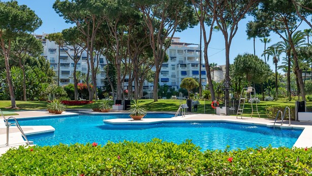 A swimming pool surrounded by greenery, a building with balconies, and a clear sky. - Apartment 'Puerto Banus2 Beachfront and Pools' with Shared Pool, Wi-Fi and Air Conditioning (Puerto Banús)