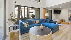 A spacious living area with a blue sofa, a round coffee table, and a large window offering a view of palm trees and a building. - Hotel Huntington (Duarte)