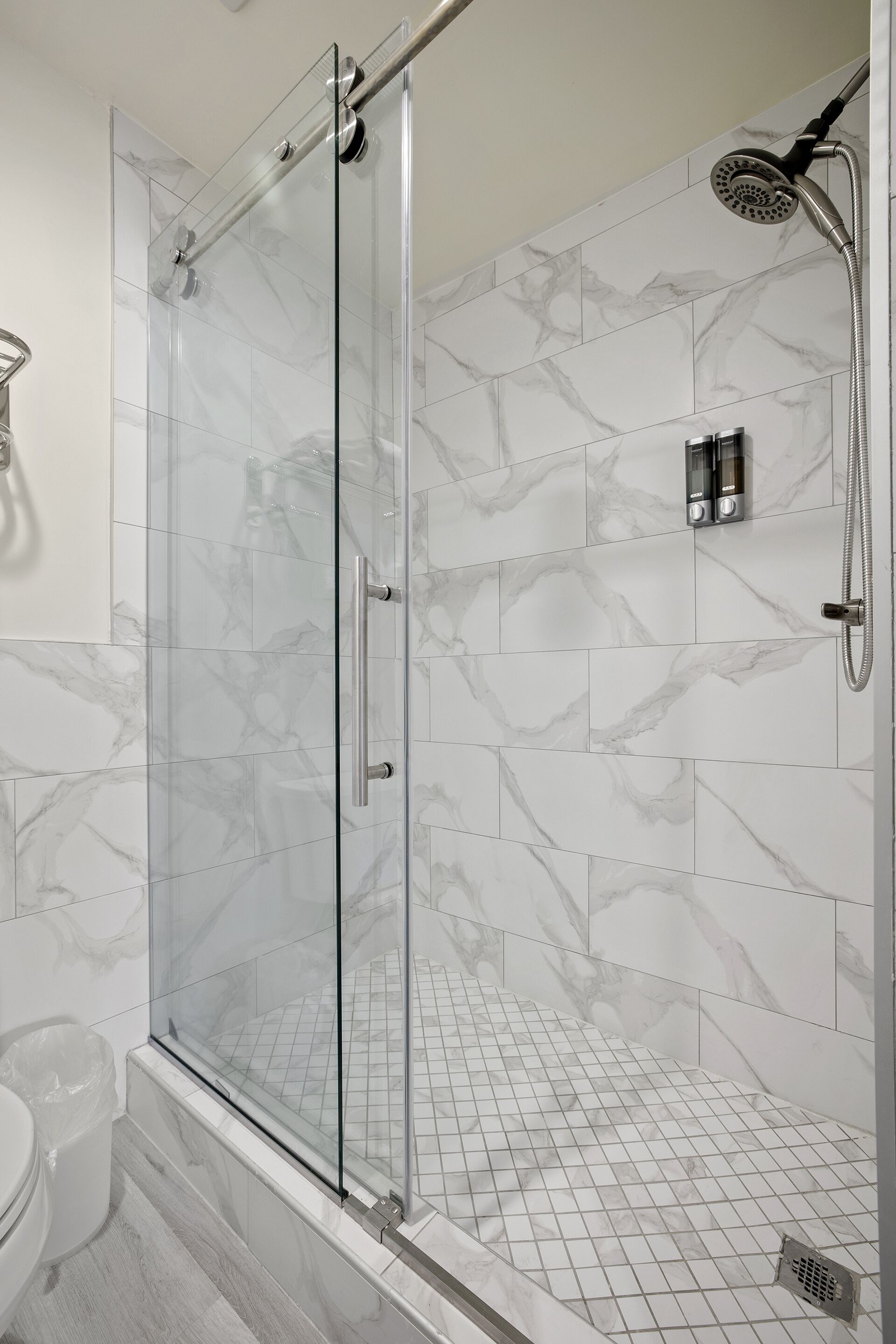 A glass-enclosed shower with a handheld showerhead, a glass door, and a tiled floor with a marble pattern.