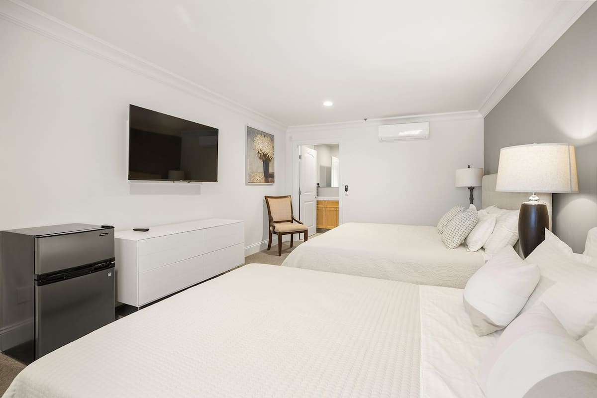 A hotel room with two beds, a desk, a chair, a TV, and a lamp.