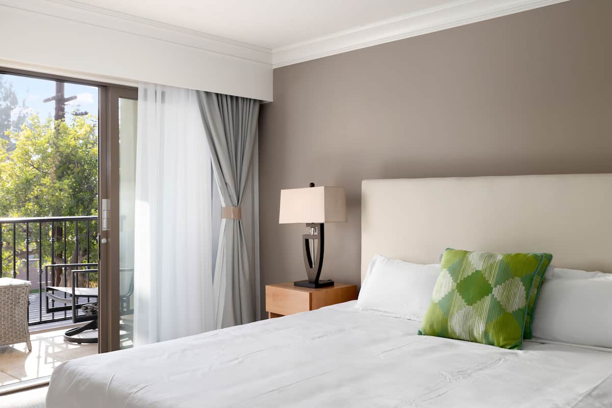 A hotel room with a bed, a nightstand with a lamp, a green pillow, and a view of a balcony with a chair and table.