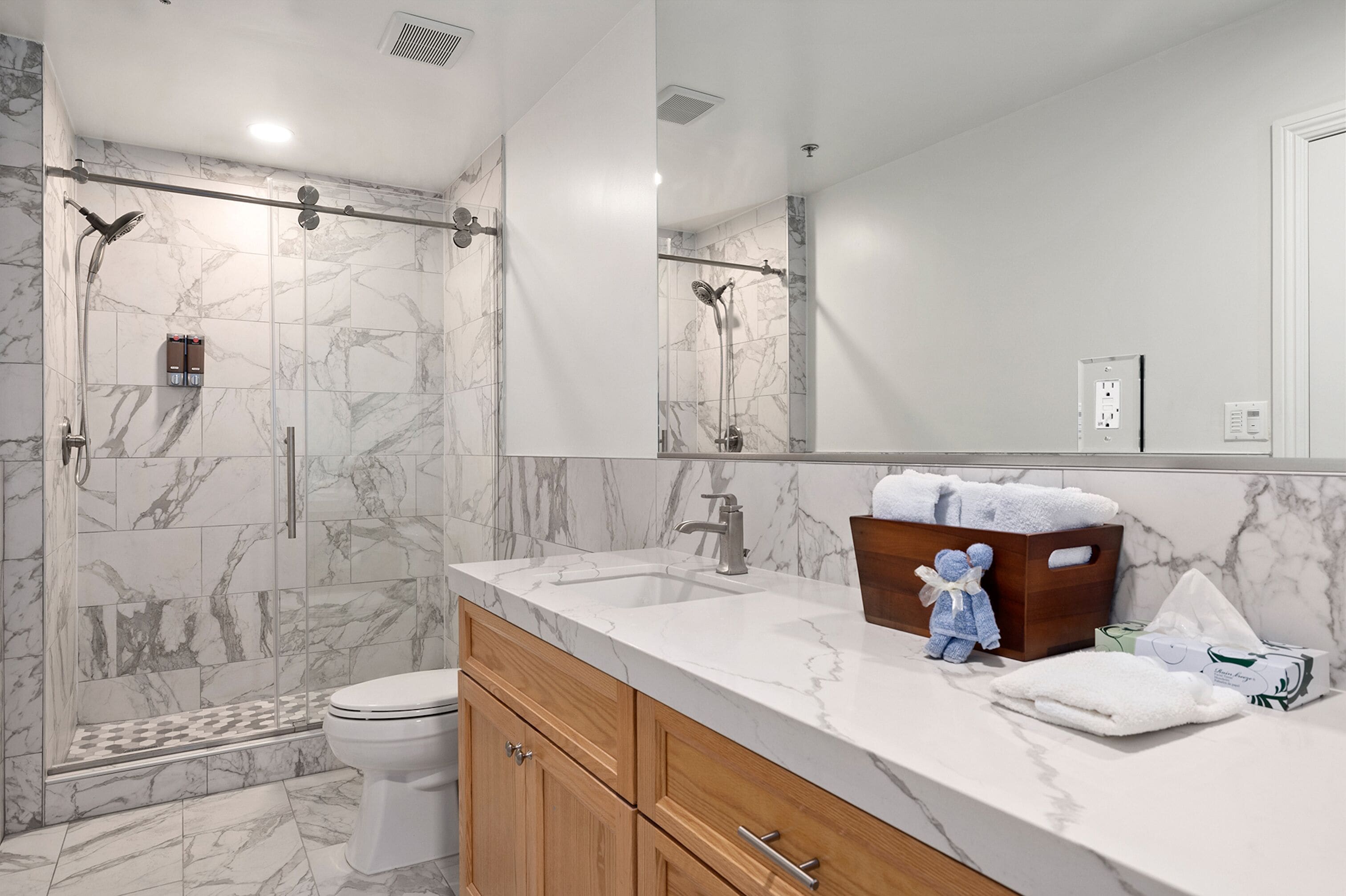 Un baño moderno con ducha de vidrio, baldosas de mármol, tocador de madera y un espejo grande.
