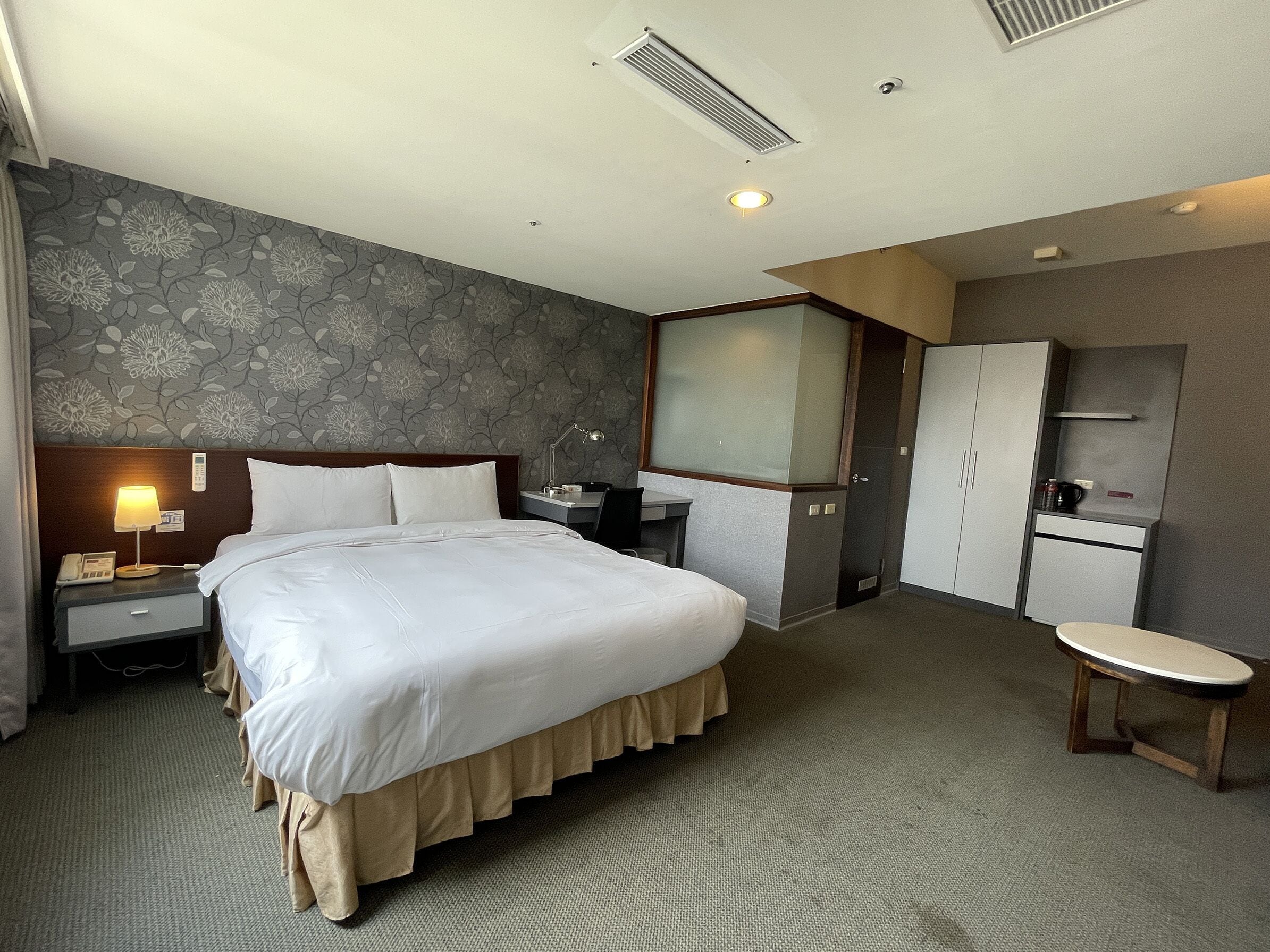 A hotel room with a large bed, a desk, a chair, a wardrobe, and a small table.