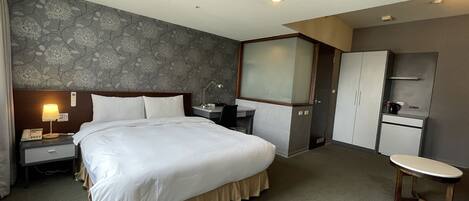 A hotel room with a large bed, a desk, a chair, a wardrobe, and a small table.