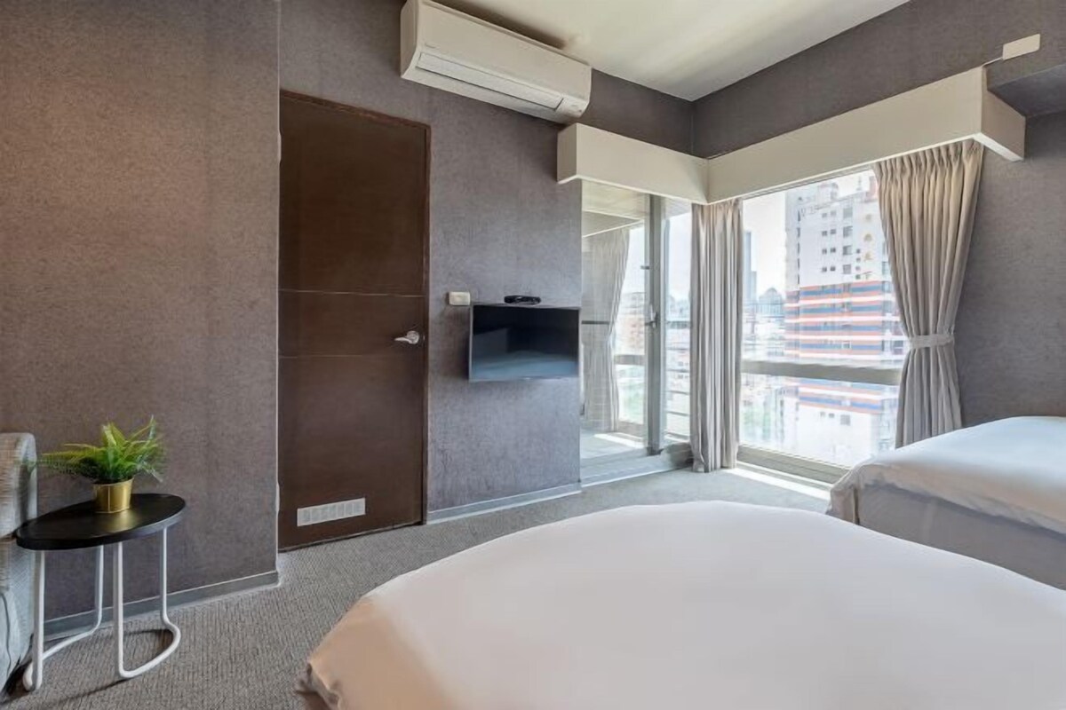Camera d'albergo moderna con un ampio letto, un tavolino laterale con una pianta, una TV a parete e una finestra con tende.