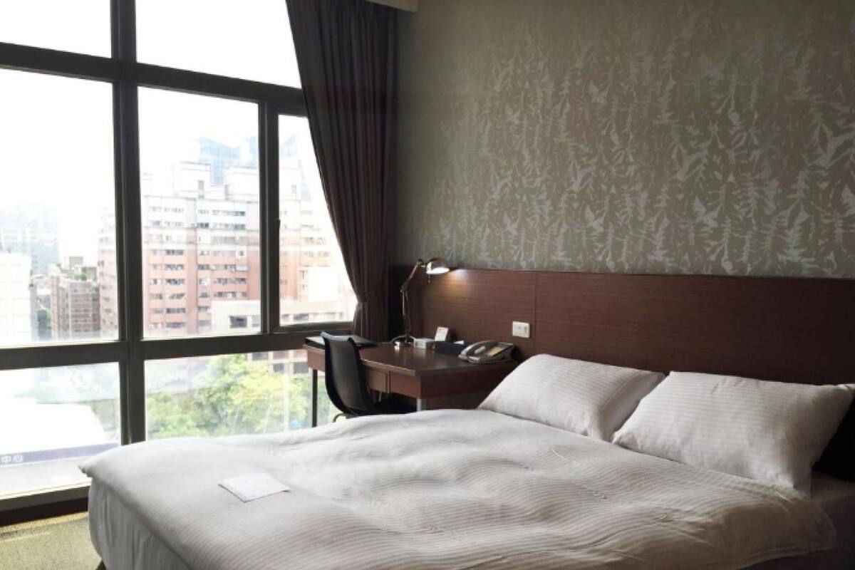 Een hotelkamer met een groot bed, een bureau met stoel, en uitzicht op de stad door grote ramen.