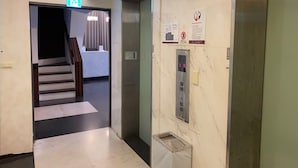 Un pasillo con una escalera que lleva a una habitación, un ascensor y un bote de basura.