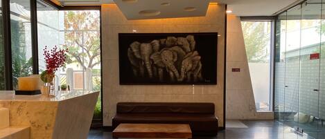 A modern lobby with a marble reception desk, a bench, a large abstract painting, and a glass wall with a view of trees.