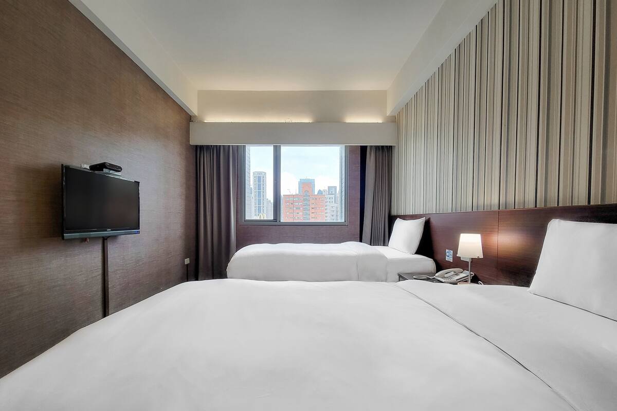 Een hotelkamer met twee bedden, een televisie, een raam met uitzicht over de stad en een lamp.