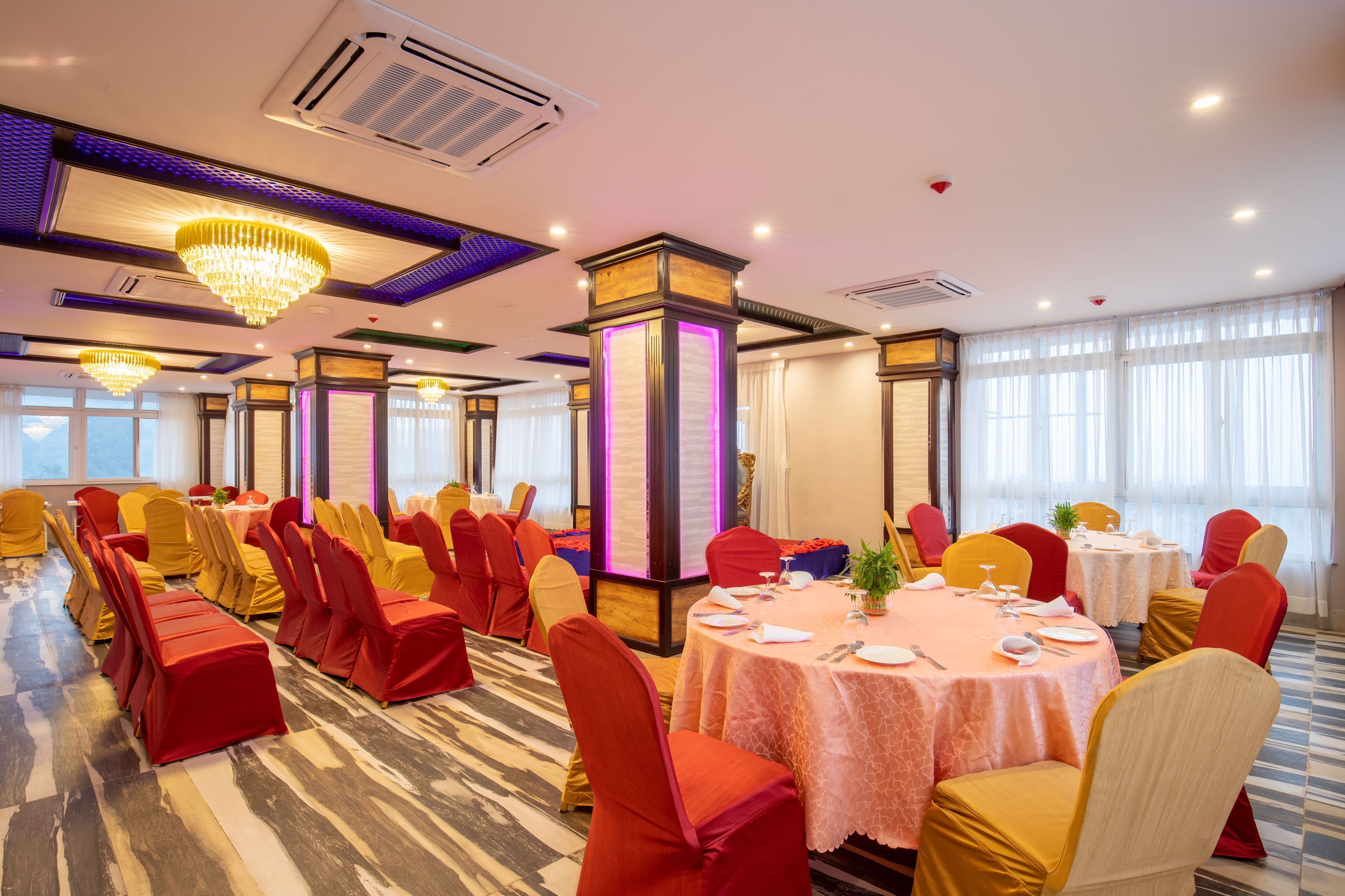 A banquet hall with round tables, red and yellow chairs, and chandeliers.