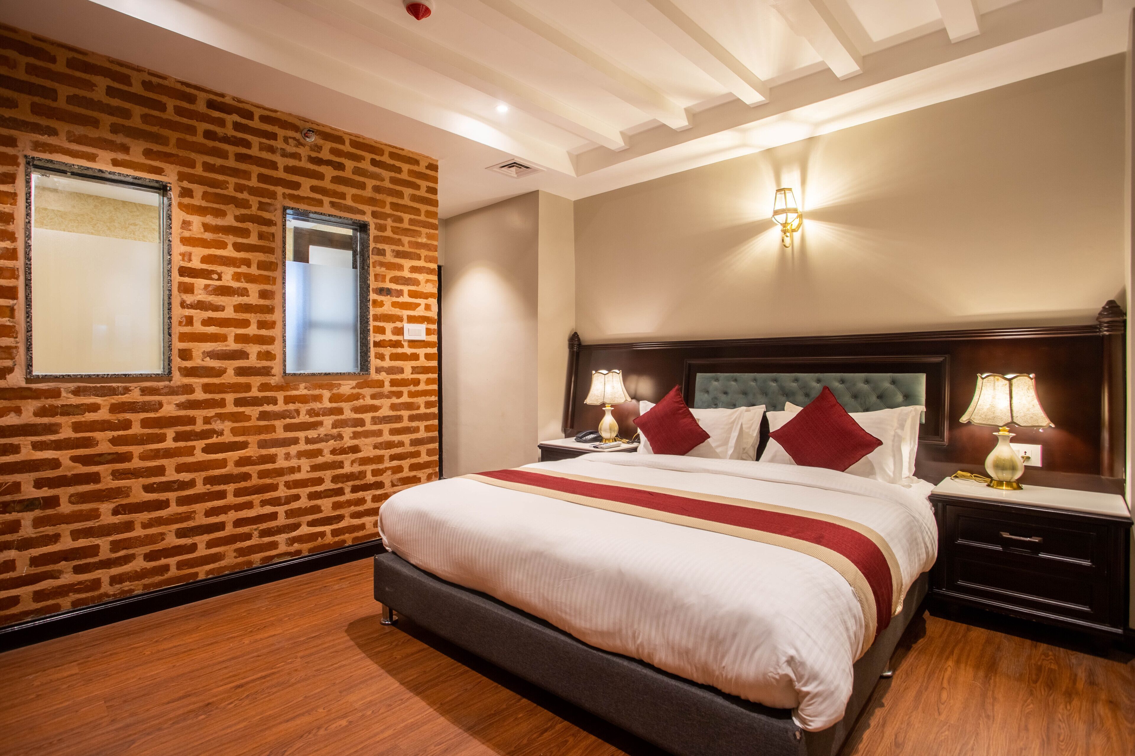 Une chambre d’hôtel avec un mur décoré d’un motif en briques, un grand lit avec une literie rouge et blanche, deux tables de chevet avec des lampes et une tête de lit au motif matelassé.