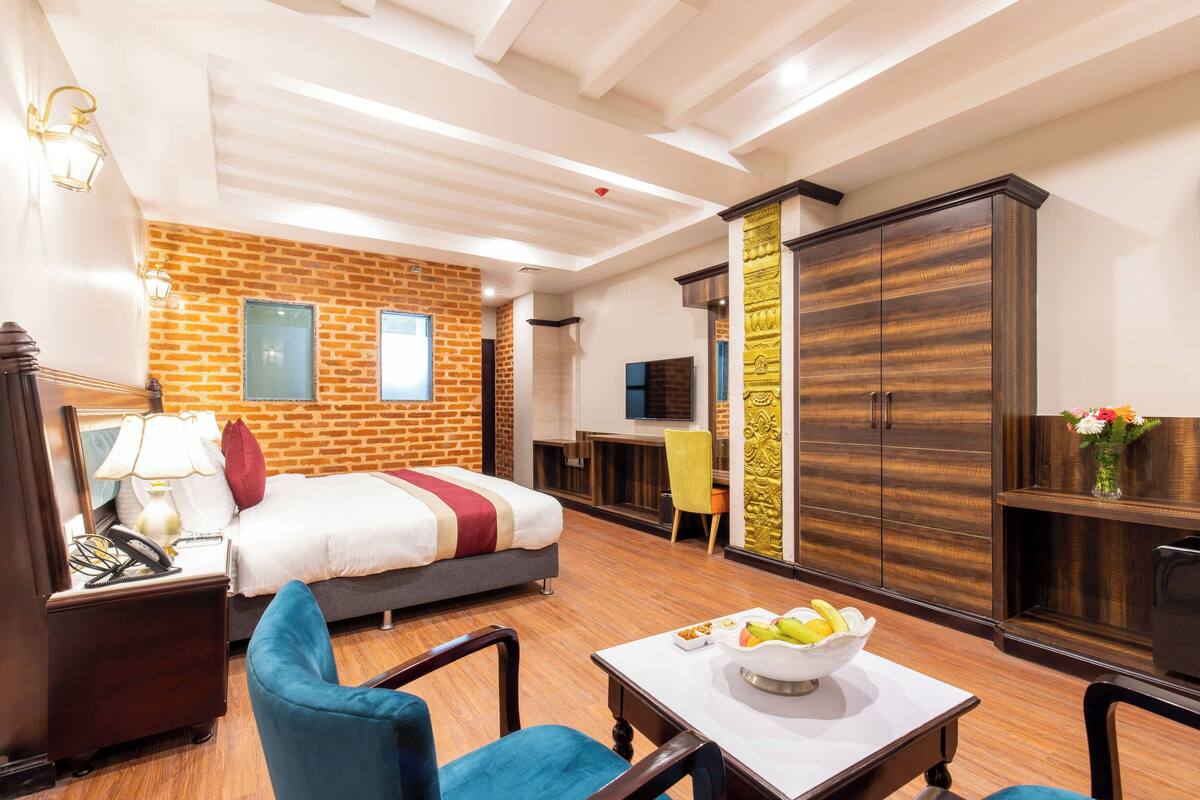 A hotel room with a brick wall, a large wardrobe, a bed, a desk, and a dining table with a bowl of fruit.