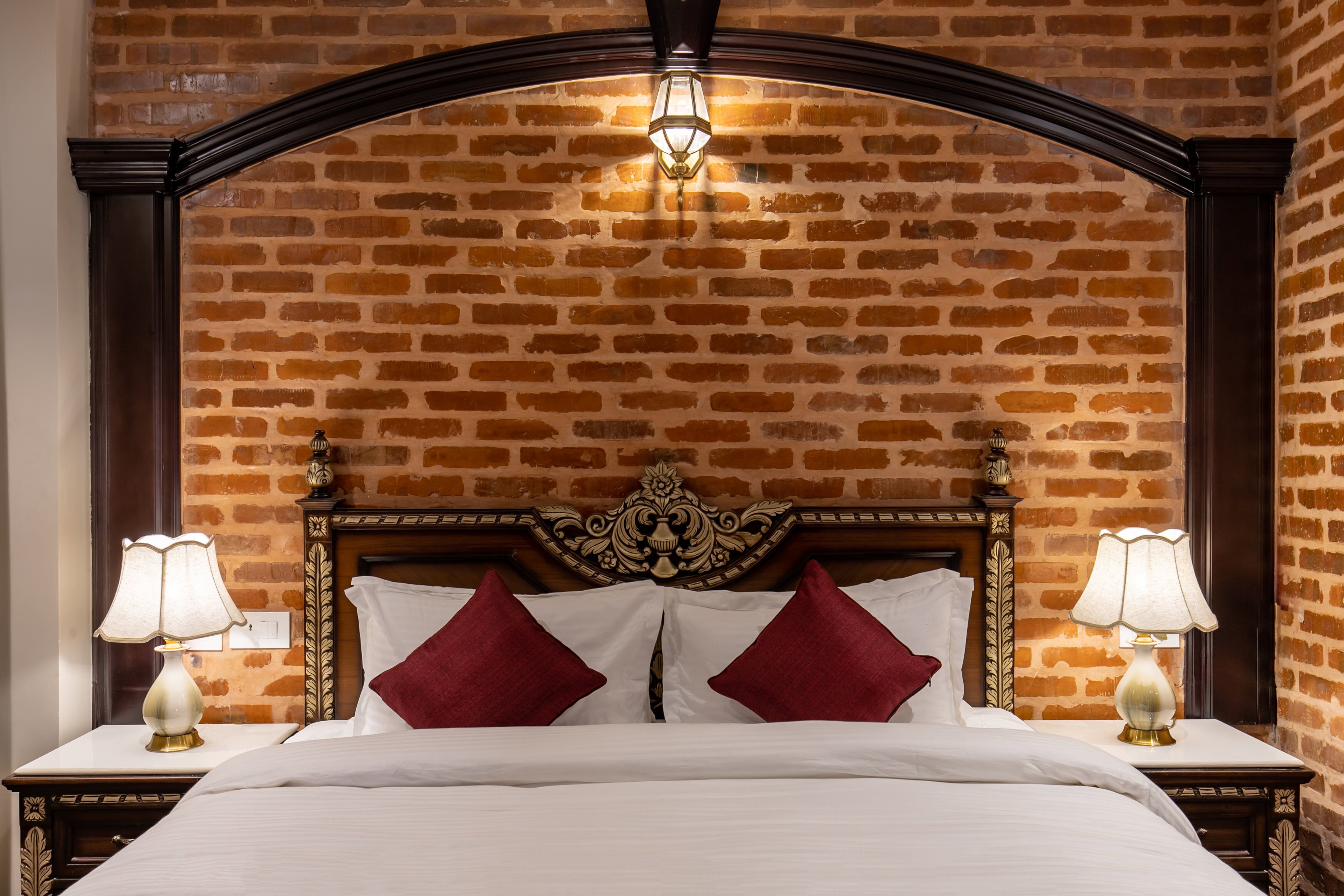 A hotel room with a brick-patterned wall, a wooden headboard, two bedside lamps, and a bed with white linens and red pillows.