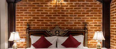 A hotel room with a brick-patterned wall, a wooden headboard, two bedside lamps, and a bed with white linens and red pillows.