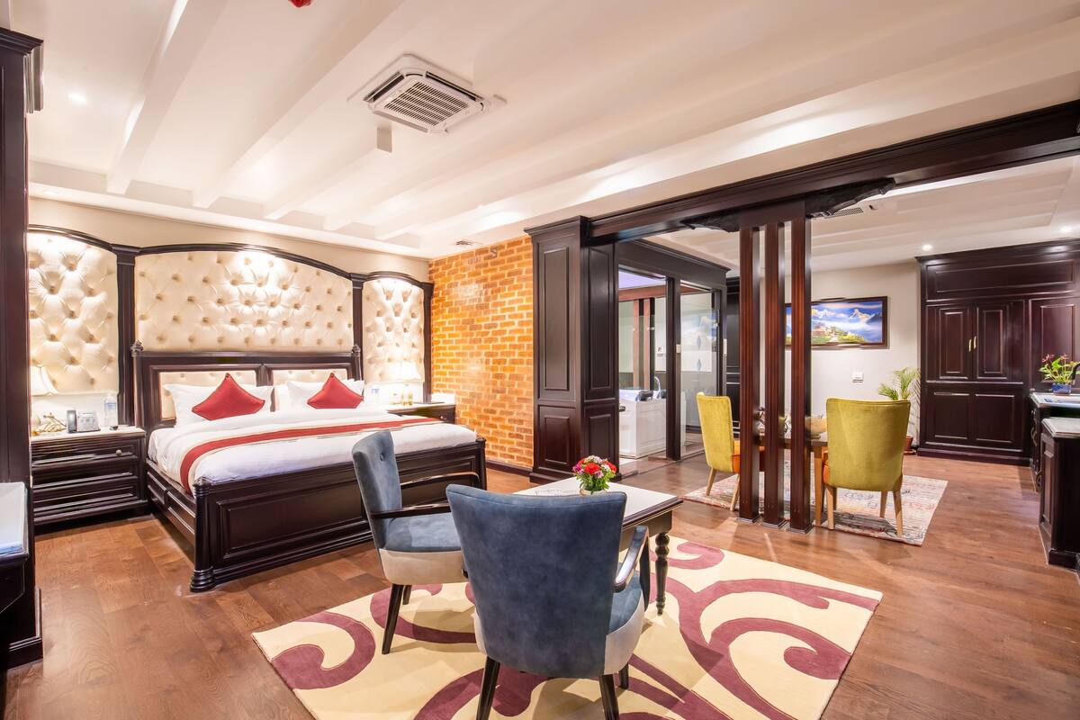 A bedroom with a large bed, a desk, two chairs, a dining table, and a kitchen area.