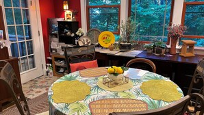 A dining area with a round table covered by a tropical leaf-patterned tablecloth, surrounded by chairs. - About Time ~~ About You  (Asheville)