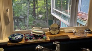 A kitchen with a window overlooking a wooded area, a sink, and various kitchen items on the counter. - About Time ~~ About You  (Asheville)
