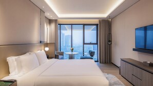 Una habitación de hotel moderna con una cama grande, un televisor, una mesita con un florero y vistas a la ciudad a través de amplios ventanales.