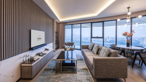 Una sala moderna con un sofá, una mesa de centro y un comedor con una mesa redonda. Se ve una vista urbana a través de ventanas amplias.