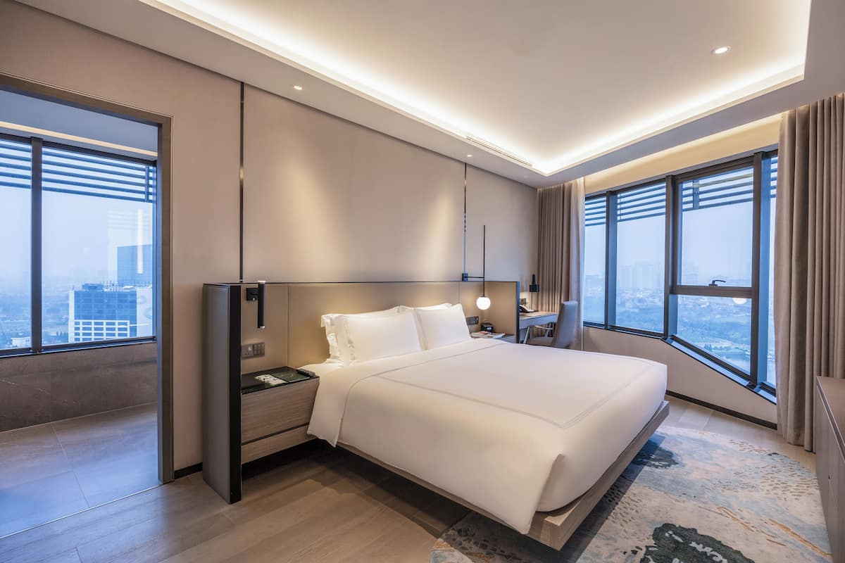 A modern hotel room with a large bed, a nightstand, a desk, and a large window offering a city view.