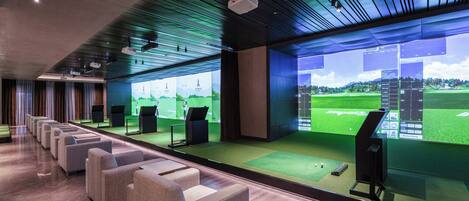 Un moderno simulador de golf en interior con filas de asientos, pantallas grandes mostrando campos de golf y un marcador digital.