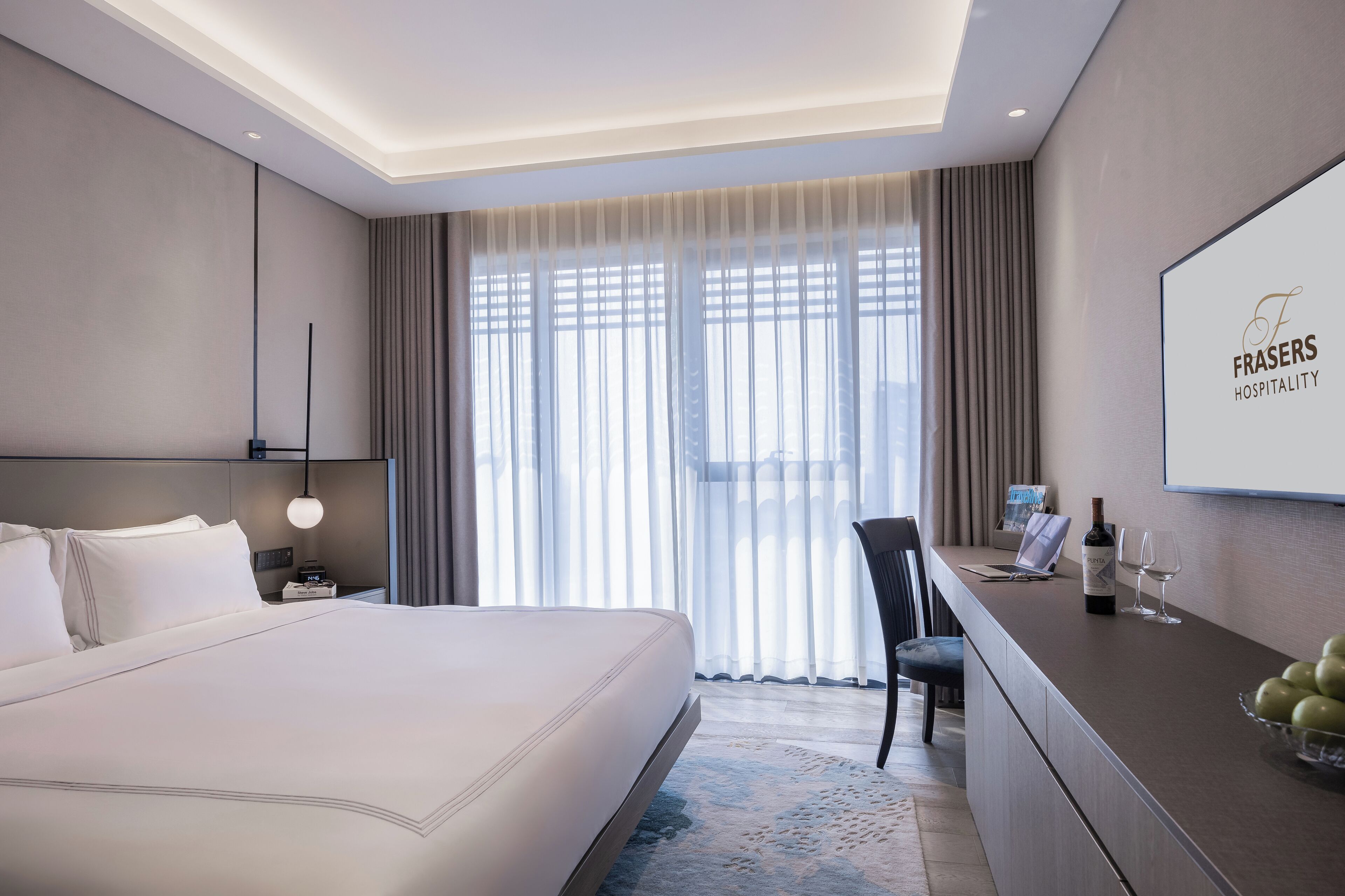 Quarto de hotel moderno com uma cama grande, uma escrivaninha com um laptop, copos de vinho e uma garrafa de vinho.
