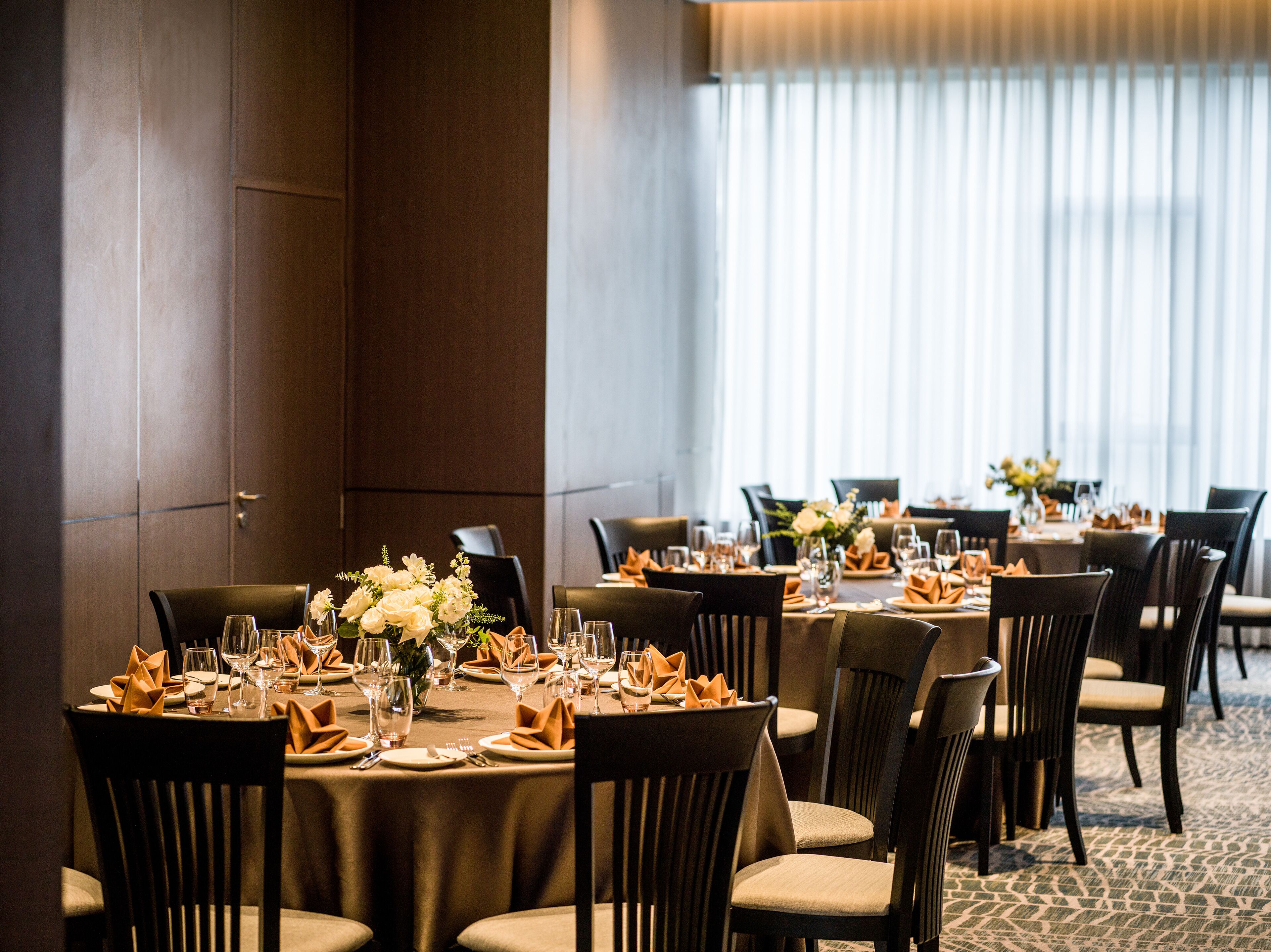 A hotel dining area with round tables set for multiple guests, featuring neatly folded napkins, wine glasses, and floral centerpieces.