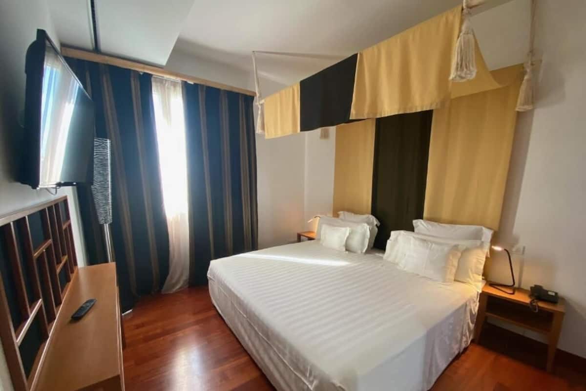 A hotel room with a large bed, a TV, a wooden desk, and a window with curtains.