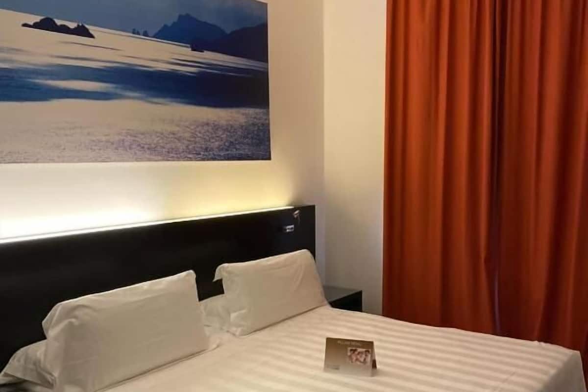 A hotel room with a bed, a nightstand, a lamp, a painting on the wall, and a window with curtains.