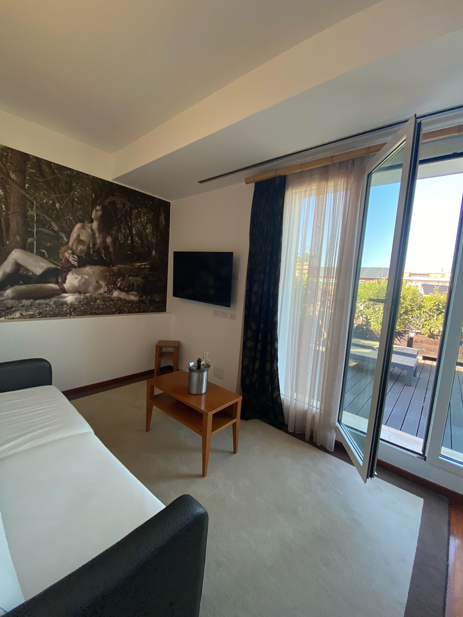 A hotel room with a bed, a desk, a chair, a painting, a TV, and a balcony with a view.