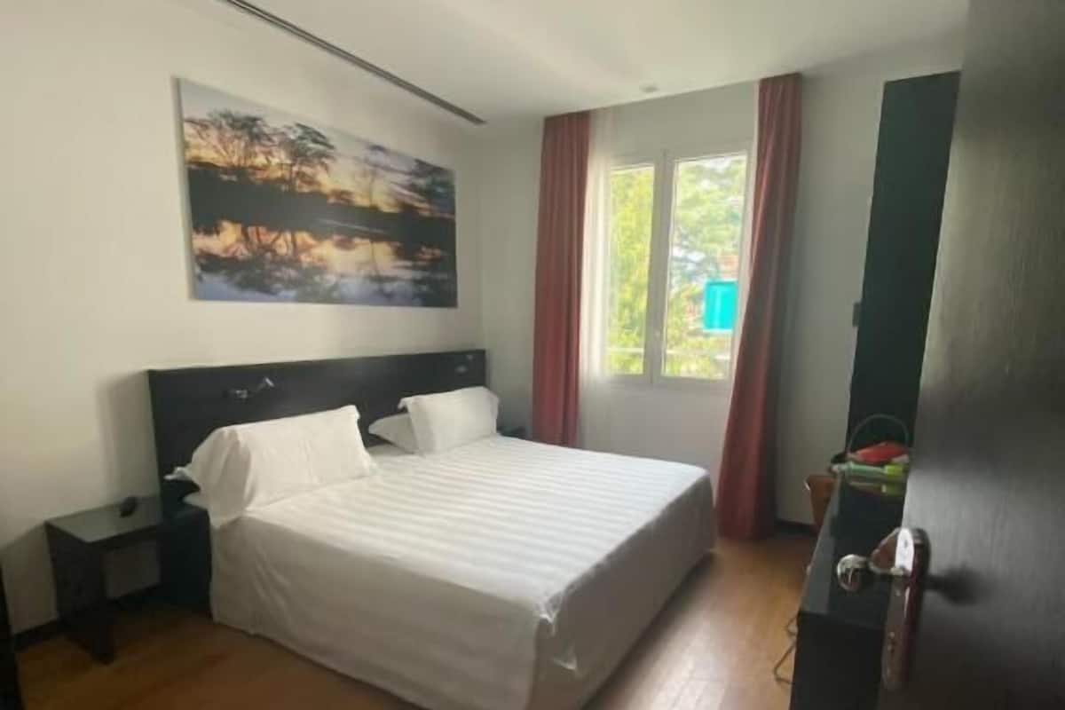 A bedroom with a bed, a nightstand, a painting on the wall, and a window with curtains.
