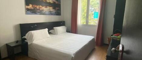 Un dormitorio con cama, mesita de noche, un cuadro en la pared y ventana con cortinas.