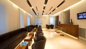 Un moderno vestíbulo de hotel con una larga recepción, zona de estar con sofás y sillones, y una pantalla de televisión en la pared.