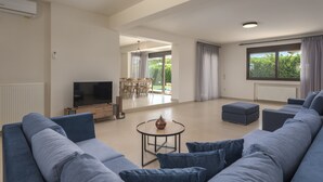 A spacious living room with a large sofa, a coffee table, and a TV. There is a dining area with a table and chairs, and a large window with curtains.